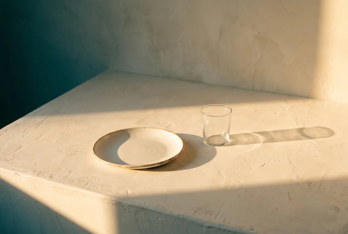 Plato y vaso vacíos sobre superficie crema con sombra alargada como reloj de sol